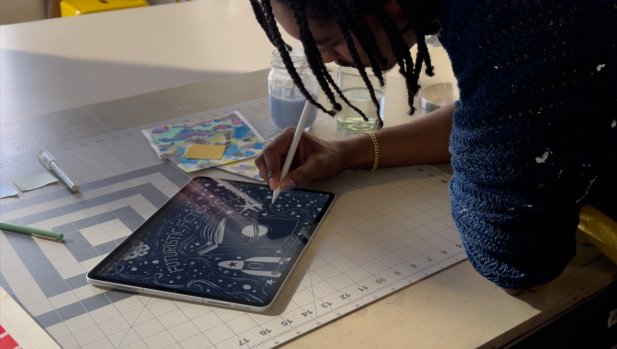 Orang berambut kepang yang mengenakan sweater biru sedang menggambar di iPad Pro dengan Apple Pencil, layar menampilkan ilustrasi langit malam dengan planet di sampul buku, pesawat luar angkasa, kupu-kupu, dan klub fiksi futuristik yang ditulis di tengah